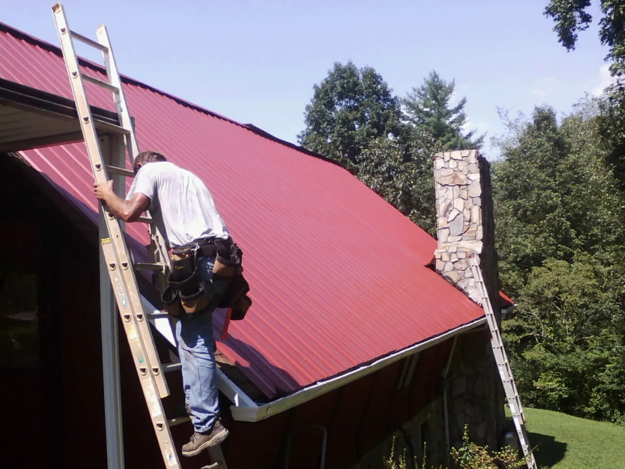 Roofer on a ladder beside a red metal roof and stone chimney — Soffit & Fascia Repair by Roofing Brothers in Dillonvale